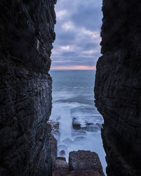 A vertical shot of the ocean under the cloudy sky visible from a gap between the rocks - Foto, imagen