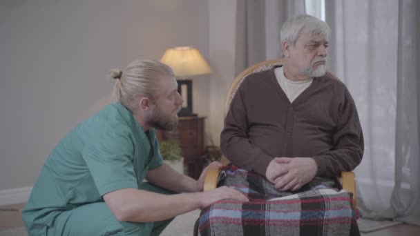 Side view of young Caucasian man talking to upset retiree sitting in armchair. Worried male nurse chatting with senior in nursing home. Support, care, help, lifestyle. - Filmati, video