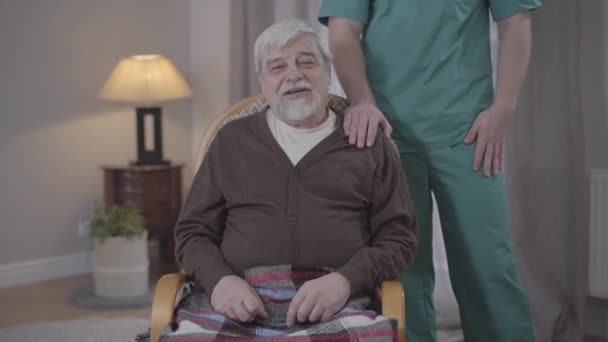 Portrait of elderly Caucasian man talking at camera as unrecognizable male nurse holding hand on his shoulder. Positive retiree spending calm evening in nursing home. Care, aging, lifestyle, help. - Кадры, видео