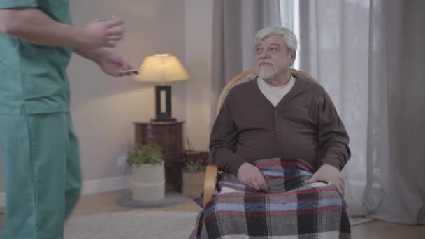 Unrecognizable nurse giving pills and glass of water to old Caucasian man sitting in armchair. Adult male volunteer taking care of senior retirees health. Help, support, lifestyle, care. - Filmagem, Vídeo