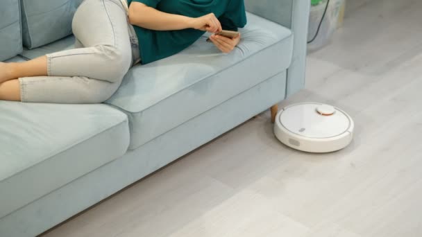 Staubsauger-Roboter. Ein elektronischer Staubsauger fährt unter den Füßen einer Frau. Frau ruht zu Hause, während ihr Staubsaugerroboter arbeitet. Moderne Heimwerkergeräte - Filmmaterial, Video