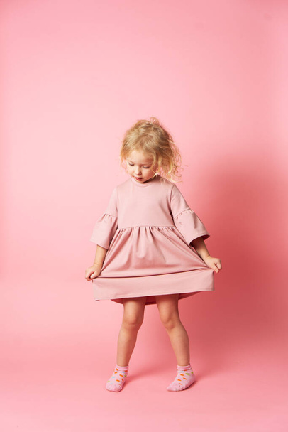 A little girl in a pink dress is spinning and demonstrating her outfit. Portrait on a pink background. - Φωτογραφία, εικόνα