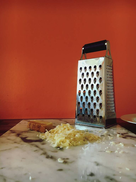 Grated cheese next to a metal grater placed on a cutting board behind an orange background - Foto, Imagen