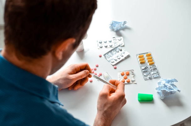 Shot of a young man holding a thermometer and many pills on a table at home. Actual problem, cold and chill, background for medical design - Foto, imagen