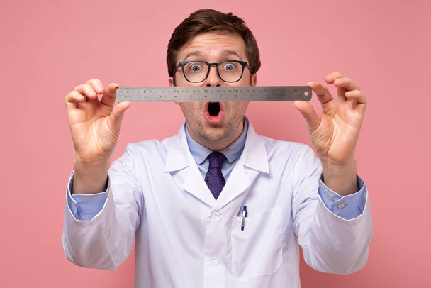 Joven médico masculino caucásico sosteniendo una regla de metal de medición con la cara conmocionada
 - Foto, imagen
