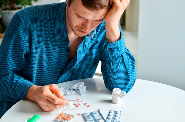 Shot of a young man holding a thermometer and many pills on a table at home. Actual problem, cold and chill, background for medical design - Foto, afbeelding
