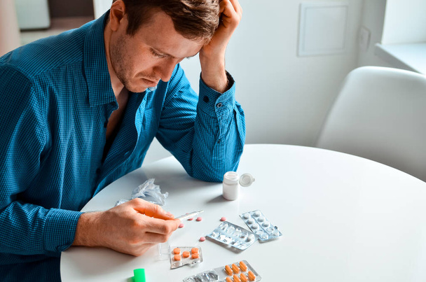 Shot of a young man holding a thermometer and many pills on a table at home. Actual problem, cold and chill, background for medical design - Foto, afbeelding
