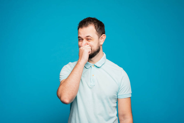 Ein Mann in einem blauen T-Shirt vor blauem Hintergrund niest lustig in seine Hand. Man kann nicht in die Hand niesen, man muss auf den Ellbogen oder in einen Schal niesen. Regelverstoß. Menschen, Krankheitskonzept - Foto, Bild