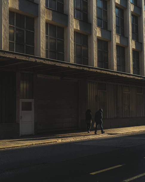 Вертикальный снимок двух человек, идущих по тротуару здания
 - Фото, изображение