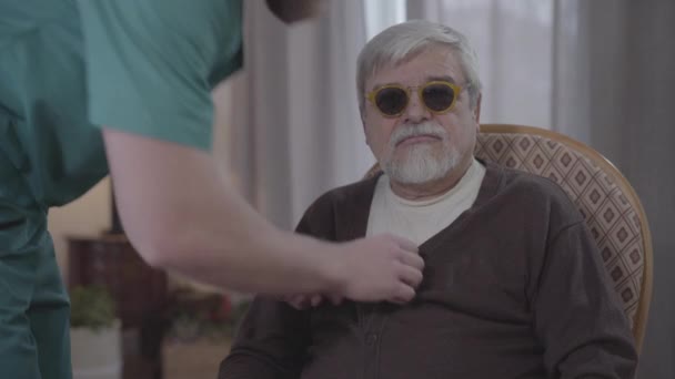Portrait of old blind Caucasian man sitting indoors as male nurse fastening button on his jacket and leaving. Adult helper taking care of mature retiree in nursing home. Lifestyle, help, support. - Кадри, відео