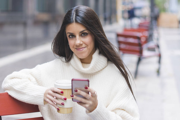 Modern glimlachend jong meisje chatten op haar mobiele telefoon met een kopje koffie zitten op een bank op straat - Foto, afbeelding