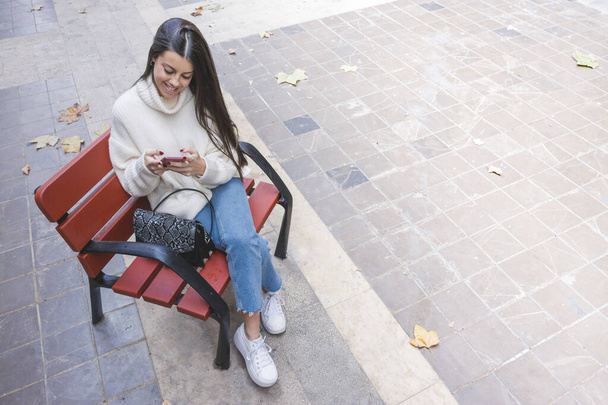 Modern mosolygós fiatal lány csevegett a mobilján egy csésze kávéval az utcán egy padon ülve - Fotó, kép