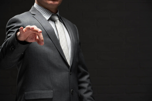zakenman demonstreert iets in zijn vingers, gekleed in grijs pak, donkere muur achtergrond - Foto, afbeelding