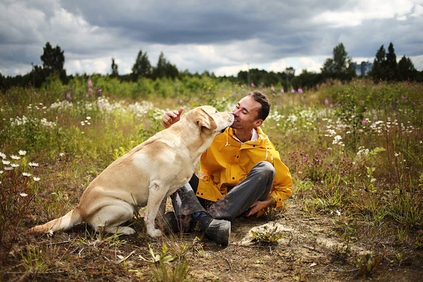 Side view of young man kissing with beige Labrador retriever in blooming meadow with cloudy sky on background - Foto, afbeelding
