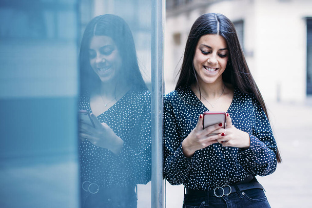 Современная и улыбающаяся молодая девушка разговаривает со своим мобильным телефоном рядом со своим отражением в витрине магазина на улице
 - Фото, изображение