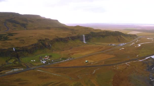 Széles légi hegyvidéki táj Izlandon, gyönyörű zöld síkság és egy kis falu a vízesés közelében. Drónfelvétel egy nagy hegyről és egy folyami deltáról. 4K - Felvétel, videó