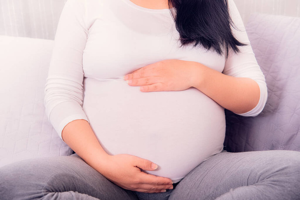 Беременная женщина, сидящая на диване и обнимающая беременного ребенка двумя руками в топике. Время материнства
.  - Фото, изображение