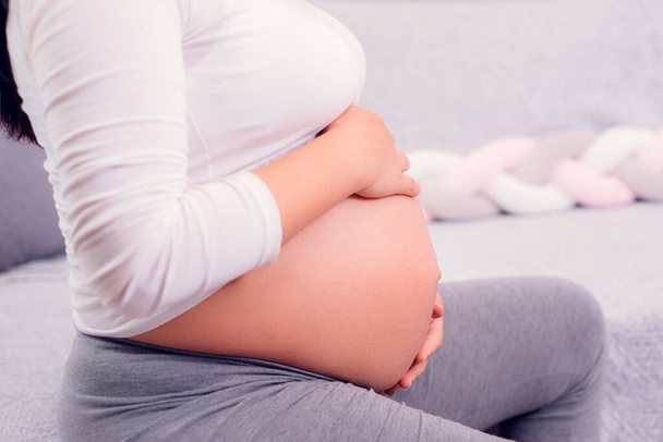 Terhes nő ül a kanapén, és két kézzel fogja a terhes kisbabáját, withe top-ban. Anyaság ideje.  - Fotó, kép