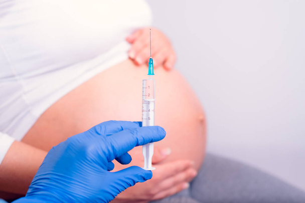 Femme enceinte ayant reçu une injection de médicament. Main avec des gants bleus tenant la seringue devant le ventre enceinte
.  - Photo, image