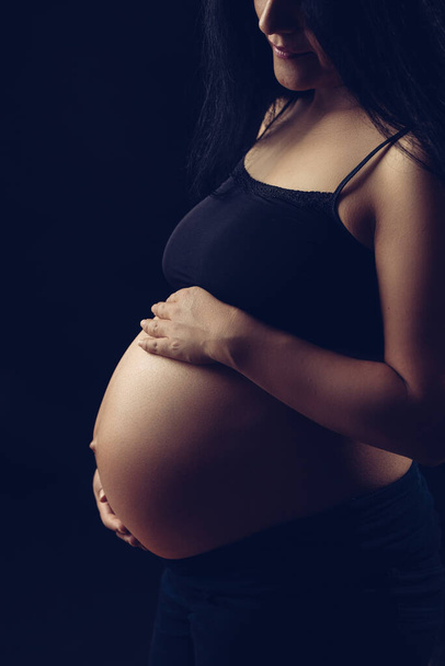 Subtle light revealing pregnant woman, holding belly with two hands. - Photo, Image
