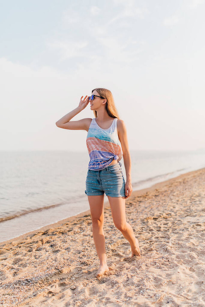 Houkutteleva nuori tyttö aurinkolasit, kävely pitkin rantaa nauttia aurinkoinen sää
. - Valokuva, kuva