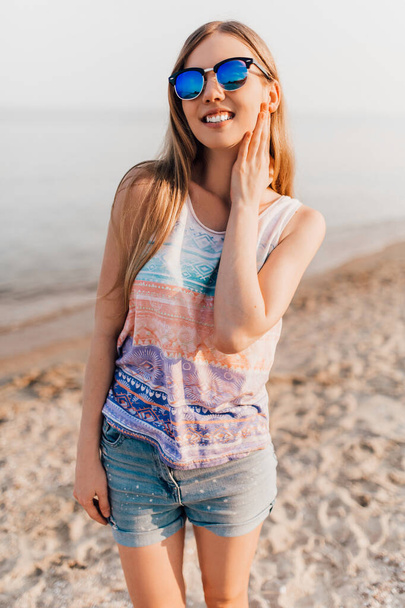 Portrait of a beautiful girl in summer clothes, laughing and looking at you while walking on the sand by the sea - Фото, изображение