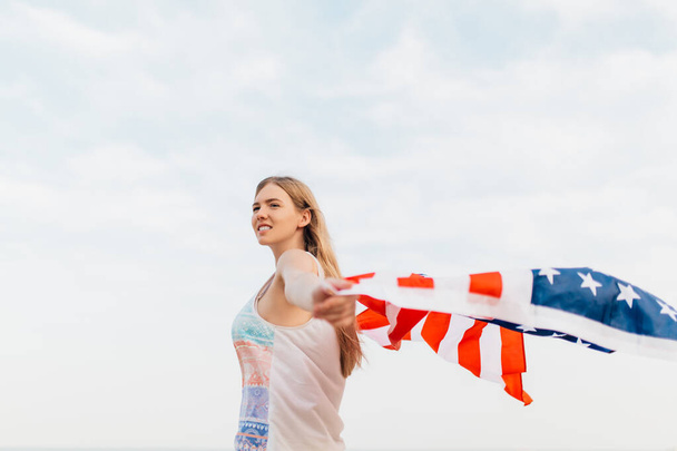 Schöne patriotische glückliche Frau mit einer amerikanischen Flagge in ihren ausgestreckten Händen, die am Strand steht - Foto, Bild