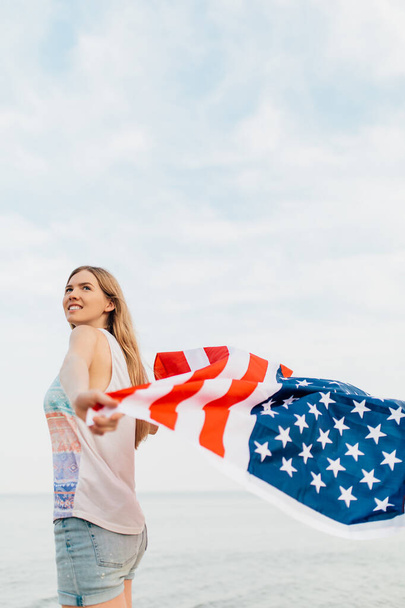 Beautiful Patriotic happy woman with an American flag in her outstretched hands standing on the beach - Foto, Imagem