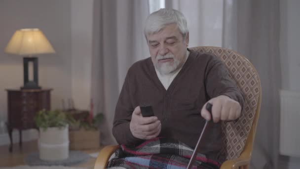Portrait of mature man with remote control arguing with TV. Grey-haired Caucasian retiree switching channels and talking at camera. Lifestyle, leisure indoors, resting. - Video, Çekim