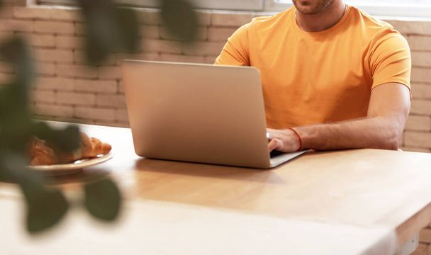 Evde sarı gömlekli bir adam var. Masada sandalyede oturup dizüstü bilgisayarla çalışıyor. Yazdırma için model. - Fotoğraf, Görsel