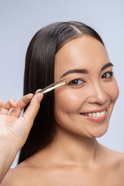 Happy Asian lady correcting eyebrows with tweezers - Zdjęcie, obraz