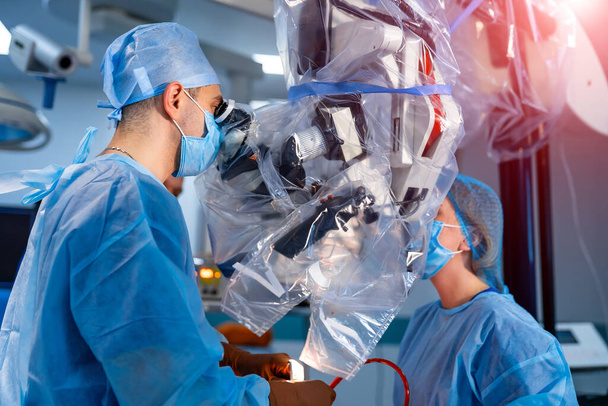 Specjalista medyczny wykonujący operacje przy użyciu nowoczesnego sprzętu. Profil chirurga w masce patrzącego w mikroskop na sali operacyjnej. - Zdjęcie, obraz