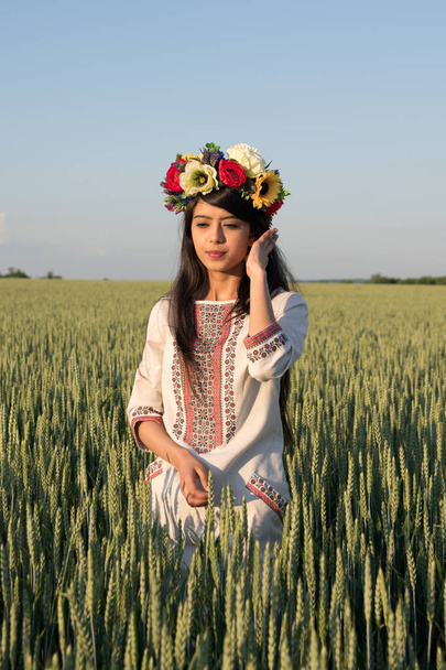 Pretty young woman wear traditional Ukrainian clothes and flower wreath walk in wheat field, beautiful ethnic girl in handmade decorated floral crown admire nature, blue sky background - Photo, Image