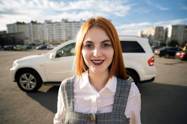 Retrato de una adolescente de moda con el pelo rojo usando vestido casual de pie afuera frente a un gran coche nuevo y caro en el cálido día de verano
. - Foto, imagen