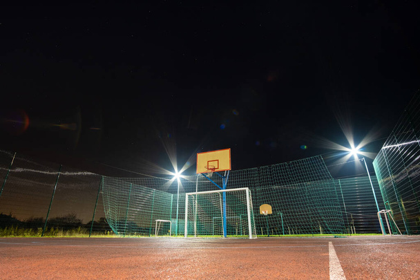Na zewnątrz mini boisko do piłki nożnej i koszykówki z bramą kulową i koszykówką otoczony wysokim ogrodzeniem ochronnym jasno oświetlone reflektorami w nocy. - Zdjęcie, obraz