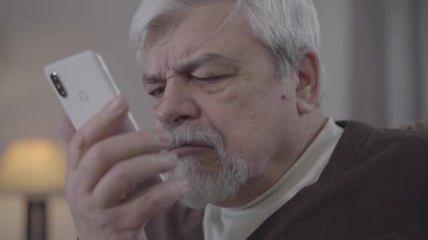 Close-up portrait of mope-eyed senior man swiping smartphone screen. Old Caucasian retiree using phone indoors. Modern technologies, lifestyle, aging, myopia. - Filmagem, Vídeo