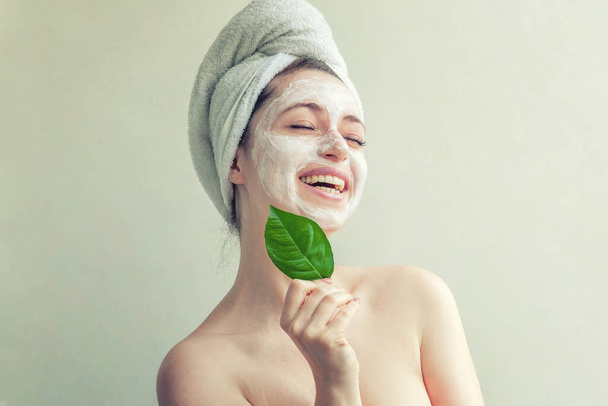 Schönheitsporträt einer Frau in Handtuch auf dem Kopf mit weißer Pflegemaske oder Creme im Gesicht und grünem Blatt in der Hand, weißer Hintergrund isoliert. Reinigung der Haut ökologisch-kosmetisches Wellness-Konzept - Foto, Bild