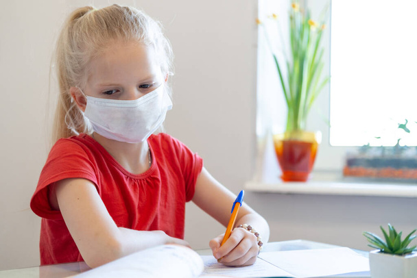 Serios sad blonde schoolgirl in medical mask stydying at home, doing school homework, writing in notebook. Reading training books on table. Back to school. Distance learning online education. - Foto, Imagem