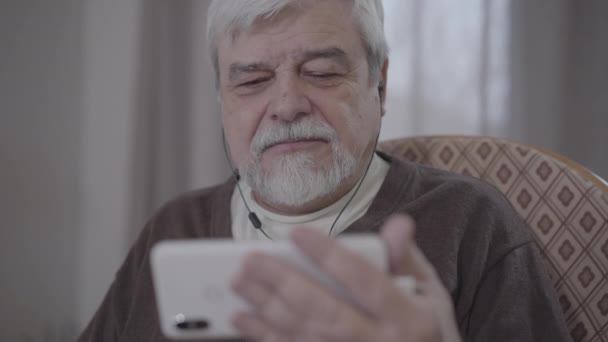 Face of brown-eyed senior man in earphones watching movie on smartphone screen. Portrait of happy retiree reacting emotionally and smiling. Lifestyle, aging, leisure, relaxation. - Video