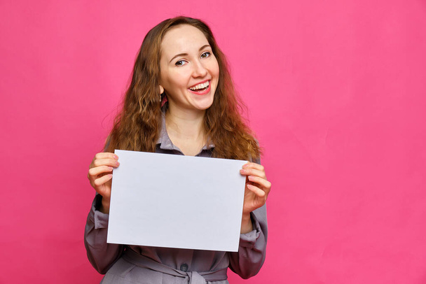 Stylish caucasian girl in a gray dress and smiles on a pink background. Holds a piece of white paper. Copy space. Close up. - 写真・画像