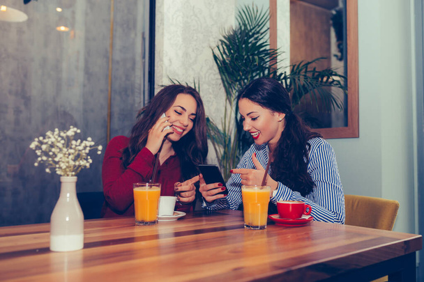 Дві молоді жінки п'ють каву, сміються і використовують смартфон у кафе
. - Фото, зображення
