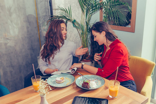 Δύο όμορφες νεαρές γυναίκες απολαμβάνουν κέικ και χυμό μαζί, ενώ διαβάζετε περιοδικό στο καφέ. - Φωτογραφία, εικόνα