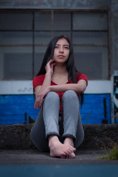 latin girl sitting on the street barefoot and dressed in red blouse and gray pants with blue background - Zdjęcie, obraz