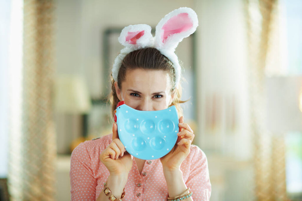 modern female in a pink blouse and easter bunny ears in the modern living room in sunny spring day hiding behind blue chicken shaped plate for eggs. - Fotó, kép