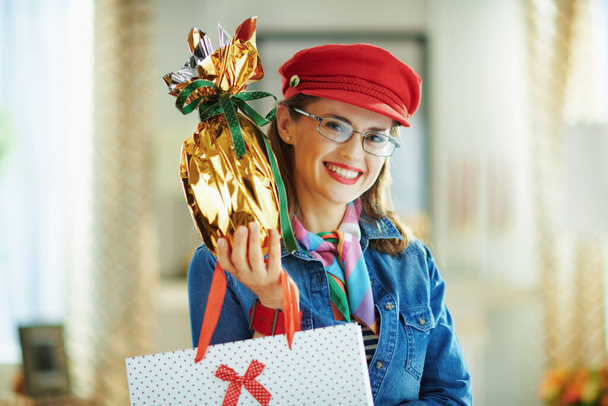 χαρούμενη μοντέρνα γυναίκα με τζιν πουκάμισο και κόκκινο καπέλο σε γυαλιά με τυλιγμένο σε χρυσό αλουμινόχαρτο μεγάλο πασχαλινό αυγό και άσπρη πουά τσάντα για ψώνια στο μοντέρνο σπίτι σε ηλιόλουστη ανοιξιάτικη μέρα. - Φωτογραφία, εικόνα