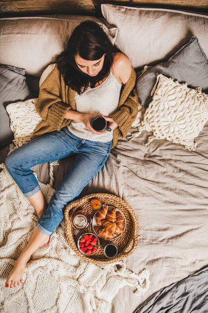 Young brunette woman in casual clothing sitting on pastel colored blankets barefoot and having breakfast in bed with fresh croissants, strawberries and tea, top view. Comfort living concept - Foto, Bild