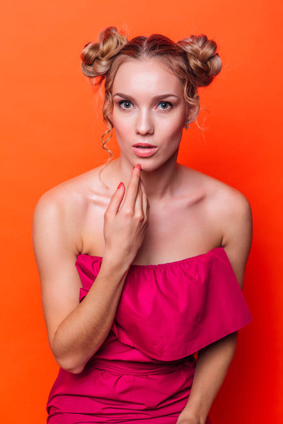 The girl in a red dress on a orange background in the studio. Blonde girl with two hair knots holding hand next to face looking to the camera and smiling. - Photo, Image