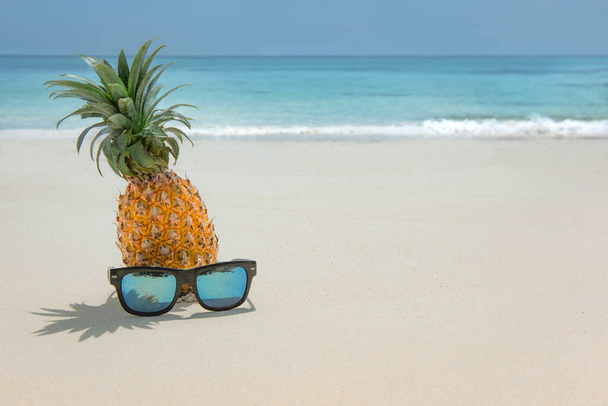 Pineapple fruit in sunglasses on sand against turquoise caribbean sea water. Tropical summer vacation concept - Photo, Image