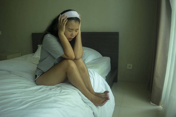 young beautiful sad and depressed Asian Korean woman in hair band and home clothes sitting on bed thoughtful feeling overwhelmed and scared about virus outbreak  - Foto, imagen