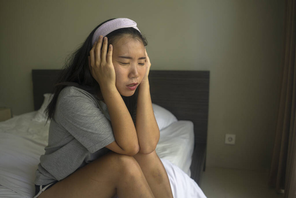 young beautiful sad and depressed Asian Chinese woman in hair band sitting on bed at home thoughtful and worried feeling overwhelmed and scared about virus outbreak and lockdown - Foto, afbeelding